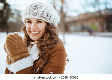 Portrait of smiling stylish middle aged woman with mittens in a knitted hat and sheepskin coat outside in the city park in winter.