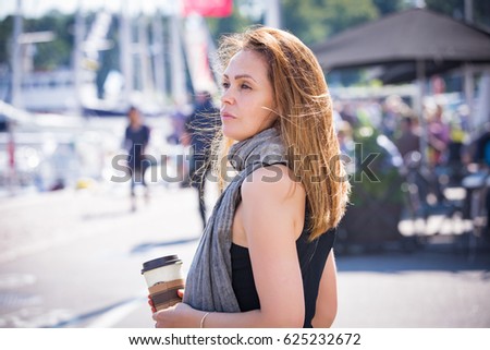 Portrait of smiling stylish middle aged woman walking along sea bay. Sailboats in harbour, seaside. Sunny summer day. Wind in the hair, drinking coffee. Vacation destination. Helsinki. Finland