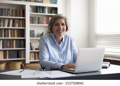 Portrait of smiling successful older 60s businesswoman, small business owner, sitting at workplace desk with computer staring at camera looking satisfied feels happy. Workday at modern office concept