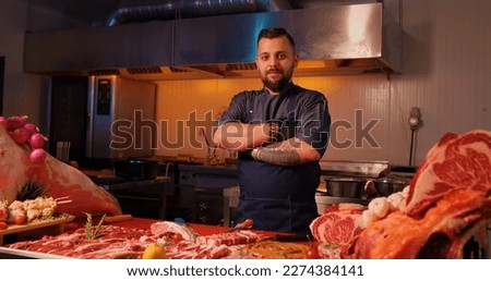 Portrait of smiling tattooed butcher in dark blue uniform at the meat manufacturing. Male worker crossing arms confident man in butchery.