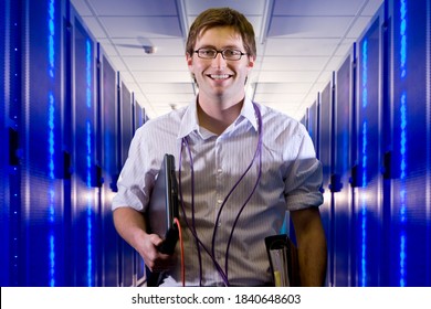 Portrait Of A Smiling IT Technician With Laptop And LAN Cables Standing In The Middle Of A Network Server Room