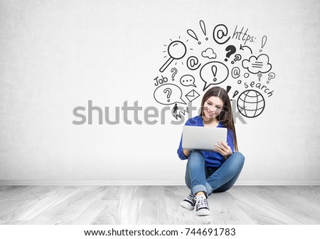 Portrait of a smiling teen girl with long dark hair wearing blue jeans and a shirt and holding her laptop while sitting on the floor near a concrete wall with an internet search sketch on it. Mock up