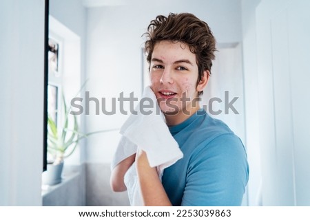 Portrait of smiling teenage boy with acne problem who takes care his face skin at home. He looking in the mirror and wipes face with towel in bathroom. Teenager skin care every day treatment process.