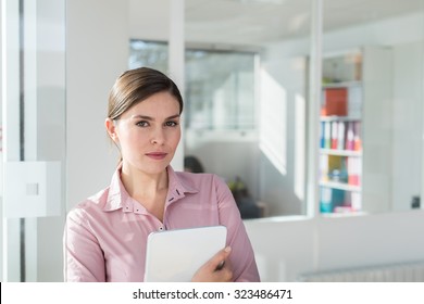 Portrait Of A Smiling Thirty Years Old Woman With Her Hair Tied And Wearing A Pink Shirt. She Is Holding A Tablet, Standing In The Hallway Of A White Office, Next To A Glass Door.