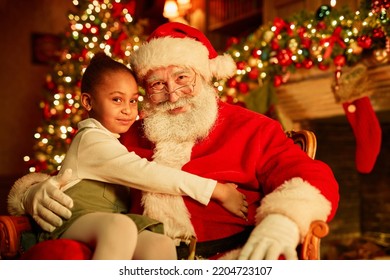 Portrait of smiling traditional Santa Claus with cute little girl sitting in his lap by Christmas tree - Shutterstock ID 2204723107