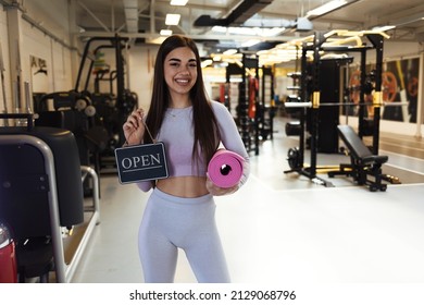 Portrait Of Smiling Trendy 20 Years Old Gym Owner Woman, Holding Mats And Showing Open Sign. Opening Fitness Centar After Covid-19 Pandemic. People And Healthcare Concept. Copy Space.