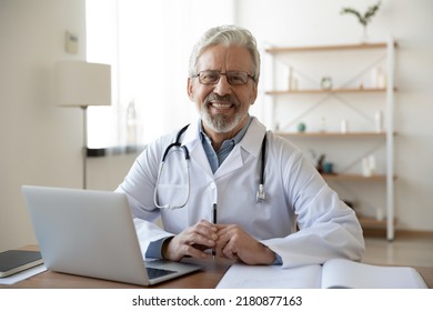 Portrait Of Smiling Trusted Middle Aged Old Doctor In Glasses And Medical White Coat Sitting At Workplace With Computer And Registry Journal, Waiting For Patient At Clinic, Medical Insurance Services.