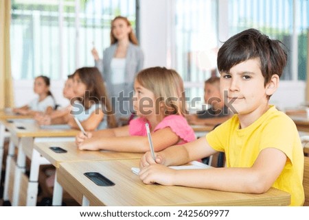 Portrait of smiling tween schoolboy sitting in classroom during lesson, writing exercises in workbook ..