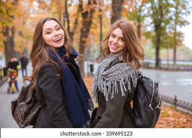 Portrait Of A Smiling Two Girlfriends Walking In Autumn Park And Looking Back At Camera