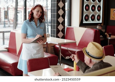 Portrait of smiling waitress in bright blue uniform taking an order from elderly gentleman in retro diner with red booths and decor featuring vinyl records - Powered by Shutterstock