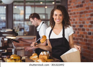 Portrait of smiling waitress putting bread roll in paper bag at coffee shop