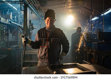 Portrait of a smiling welder posing in workshop while his colleague in the background is working with grinder. They are both in work uniforms and have protective helmets on their heads.