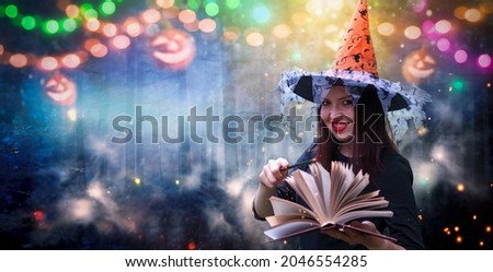 Portrait of smiling witch with open magic book of spells and wand on festive forest background. Beautiful young woman in witches orange hat conjuring,making witchcraft.Halloween party art design