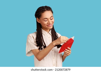 Portrait Of Smiling Woman With Black Dreadlocks Standing Open Red Envelope With Congratulations, Reading Romantic Letter, Wearing White Shirt. Indoor Studio Shot Isolated On Blue Background.