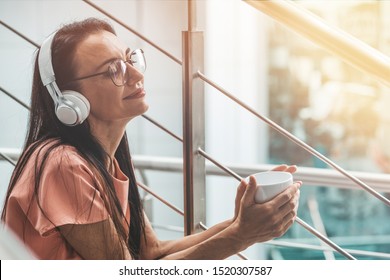 Portrait of smiling woman in casual wear and glasses sitting on the stairs, drinking coffee or tea while listening to music via headphones. Horizontal shot. Side view - Powered by Shutterstock