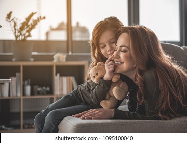 Portrait Of Smiling Woman And Child Lying On Sofa. Little Girl Is Touching Her Mom Nose With Her Finger Holding Teddy Bear. Cute Family Is Happy And Looking Perfect