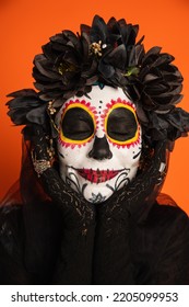 Portrait Of Smiling Woman With Closed Eyes And Creepy Halloween Makeup Holding Hands Near Face Isolated On Orange