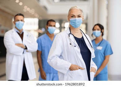 Portrait Of Smiling Woman Doctor Looking At Camera And Standing In Hospital With Team In Background Wearing Protective Face Mask During Covid-19 Pandemic. Healthcare Workers Team During Corona Virus.