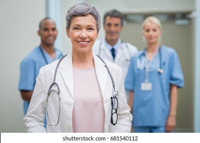 Portrait Of Smiling Woman Doctor Standing In Hospital With Team In Background. Senior Female Doctor In Front Of Her Medical Staff. Team Of Medical Professionals Looking At Camera.