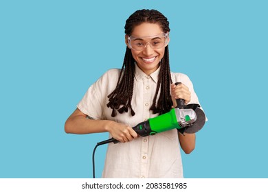 Portrait of smiling woman with dreadlocks holding grinder saw, looking at camera with positive emotions, wearing white shirt and protective glasses. Indoor studio shot isolated on blue background.