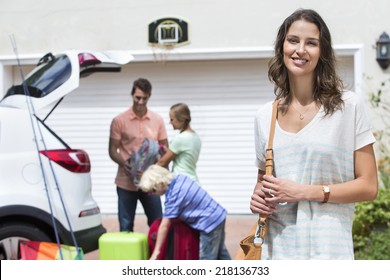 Portrait Of Smiling Woman In Driveway With Family Packing Car In Background