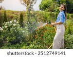 Portrait of smiling woman gardener watering plants with hose pipe in summer garden. Taking care of blooming flowers.