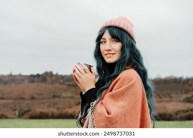 Portrait of smiling Woman in hat wrapped in Shawl Poncho drinking hot drink from thermos cup enjoying autumn landscape view. Feeling harmony, relax, personal fulfillment. Local travel lifestyle. - Powered by Shutterstock