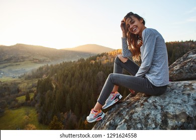 Portrait Of Smiling Woman Hiker Sitting On Edge Of Cliff Against Background Of Sunrise