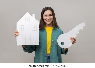 Portrait Of Smiling Woman Holding Big Key And Paper House, Looking At Camera With Smile, Purchase Of Real Estate, Wearing Casual Style Jacket. Indoor Studio Shot Isolated On Gray Background.