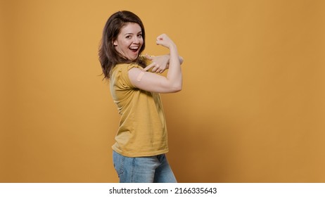 Portrait Of Smiling Woman Lifting Sleeve And Showing Band Aid After Covid Or Flu Vaccine Flexing Arm Biceps In Studio. Proud Person Revealing Bandage Covering Immunization Spot For Medical Awareness.