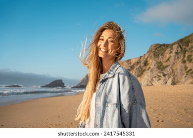 Portrait of smiling woman with long blond hair on sandy beach. Happy female in denim shirt with sea and mountain in background. Lady enjoying vacation against blue sky. - Powered by Shutterstock