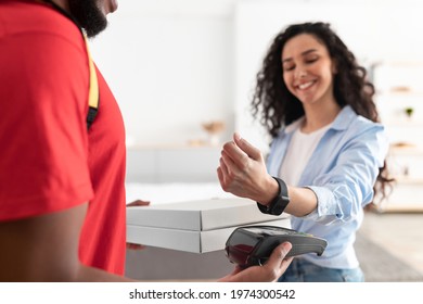 Portrait of smiling woman making payment through wireless modern bank terminal. Consumer paying using smart watch and NFC technology to courier for package delivery. Modern Futuristic Technology