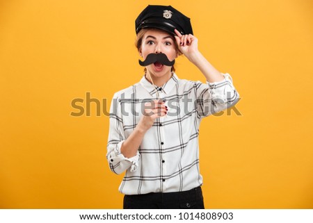 Portrait of smiling woman in plaid shirt and police cap holding paper moustache at her face isolated over yellow background