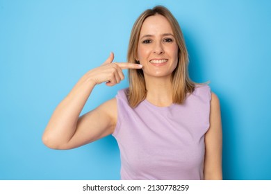 Portrait of smiling woman pointing at her nice teeth and mouth isolated on blue background. Dental concept.