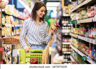 Portrait Of Smiling Woman With Shopping Cart In Supermarket Buying Groceries Food Walking Along The Aisle And Shelves In Grocery Store, Holding Glass Jar Of Sauce, Choosing Healthy Products In Mall - Shutterstock ID 1873973239
