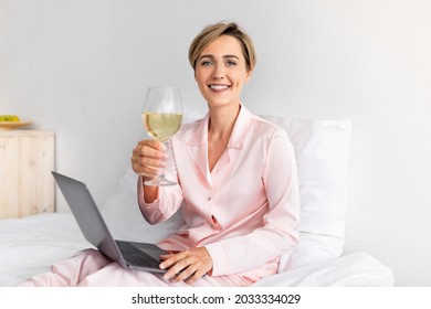 Portrait of smiling woman sitting on bed using laptop and enjoying wine after beauty care. Woman wearing pink silk sleepwear resting with computer at home, looking and toasting at camera - Powered by Shutterstock