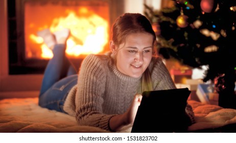 Portrait Of Smiling Woman In Sweater Lying By The Fireplace Under Christmas Tree And Using Digital Tablet