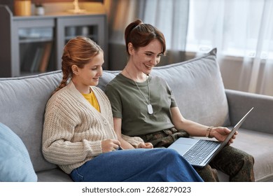 Portrait of smiling woman wearing military uniform using laptop while sitting on sofa with teen daughter - Powered by Shutterstock