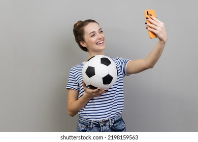 Portrait Of Smiling Woman Wearing Striped T-shirt Making Selfie, Having Video Call Or Broadcasting Livestream With Ball In Hand. Indoor Studio Shot Isolated On Gray Background.