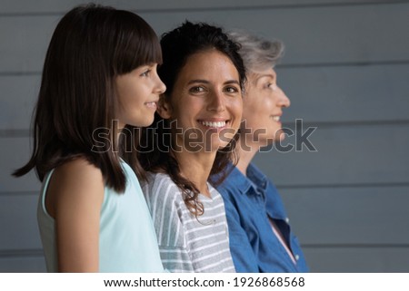 Portrait of smiling young 30s Latino woman with little daughter and elderly grandmother pose on grey background. Three generations of women in row, adult grownup female with child and grandparent.