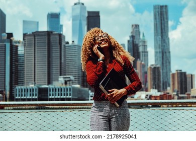 Portrait Smiling Young Adult Entrepreneur Millennial Woman With Eyeglasses And Afro Hair Talking On A Phone Call Outdoors With Manhattan New York City Skyline Skyscraper Behind Hudson River