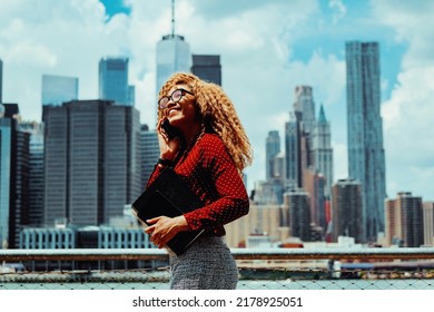 Portrait Smiling Young Adult Entrepreneur Millennial Woman With Eyeglasses And Afro Hair Talking On A Phone Call Outdoors With Manhattan New York City Skyline Skyscraper Behind Hudson River