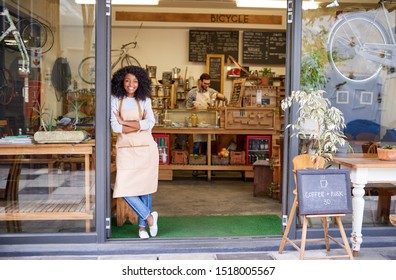 Portrait of a smiling young African American barista leaning with her arms crossed against the entrance of a trendy cafe 