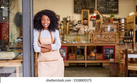 Porträt einer lächelnden jungen afroamerikanischen Barista, die sich mit den Armen an der Tür eines trendigen Cafés befindet 