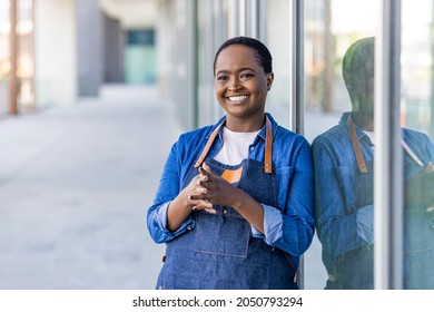 Portrait of a smiling young African American barista leaning with her arms crossed on the door of a trendy cafe. Portrait of a happy waitress standing at restaurant entrance.  - Powered by Shutterstock