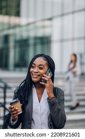 Portrait Of A Smiling Young African American Business Woman Talking On A Mobile Phone And Holding A Coffee Takeaway. Successful Businesswoman Using Smartphone And Standing On A Stairs Outside.