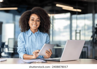 Portrait of a smiling young African American woman sitting at a desk in the office, holding a tablet and smiling at the camera. - Powered by Shutterstock