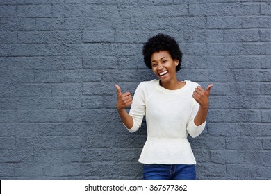 Portrait of smiling young african american woman gesturing thumbs up sign with both her hands against gray wall
