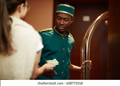 Portrait Of Smiling Young African Boy Working As Bellhop In Luxury Hotel, Getting Tip From Woman Guest For Delivering Luggage To Room