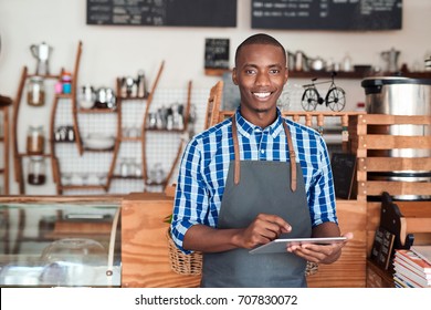 Portrait of a smiling young African entrepreneur in an apron leaning against the counter of his trendy cafe using a digital tablet - Powered by Shutterstock